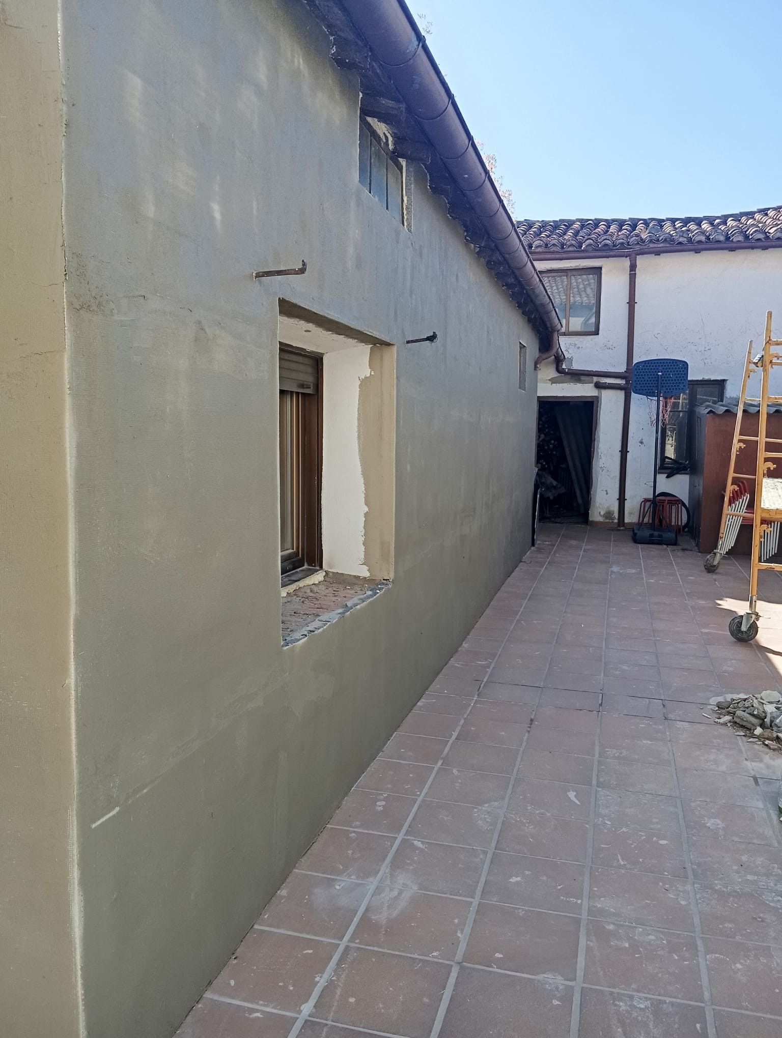 Patio estrecho pavimentado con baldosas, entre paredes claras, con una ventanita y un pasadizo abierto al fondo.