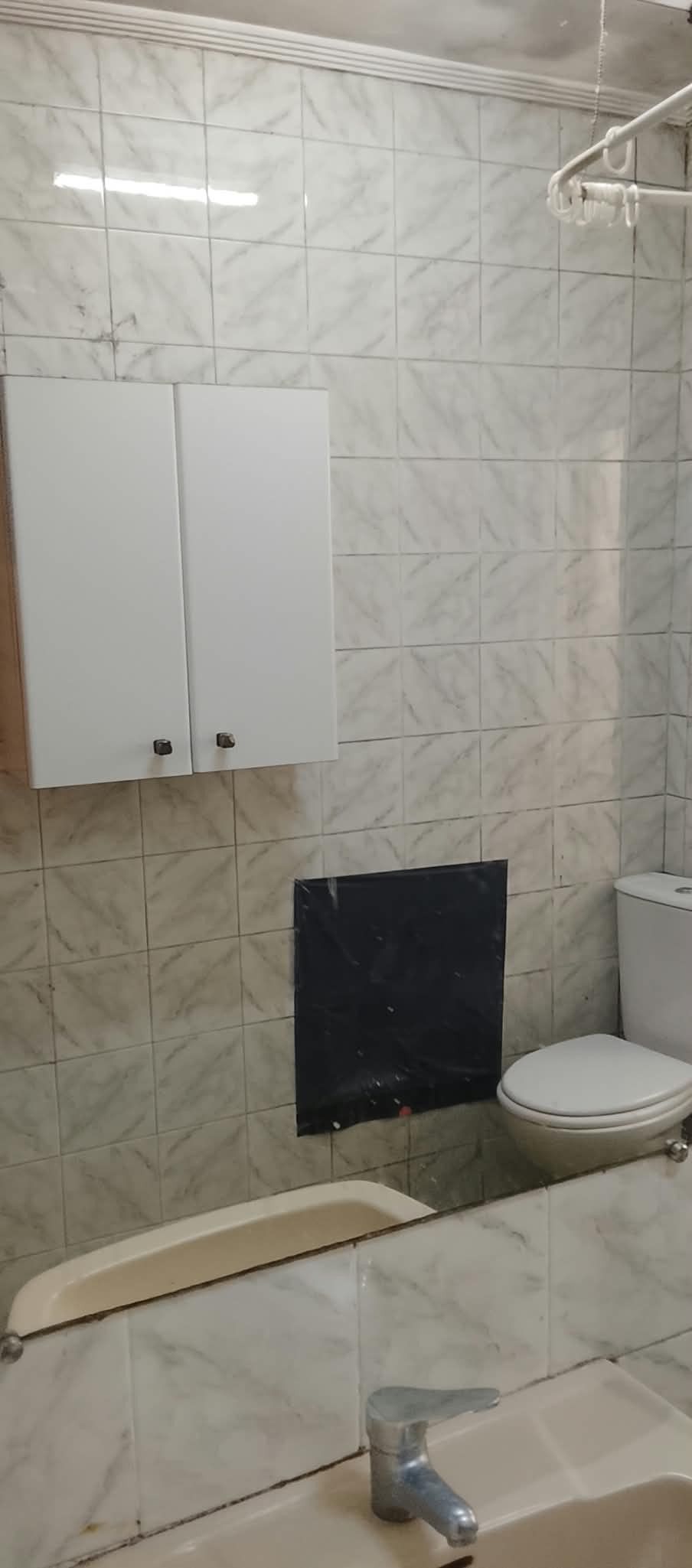 Baño alicatado con un armario de pared, lavabo, inodoro y un hueco o panel negro en la pared.