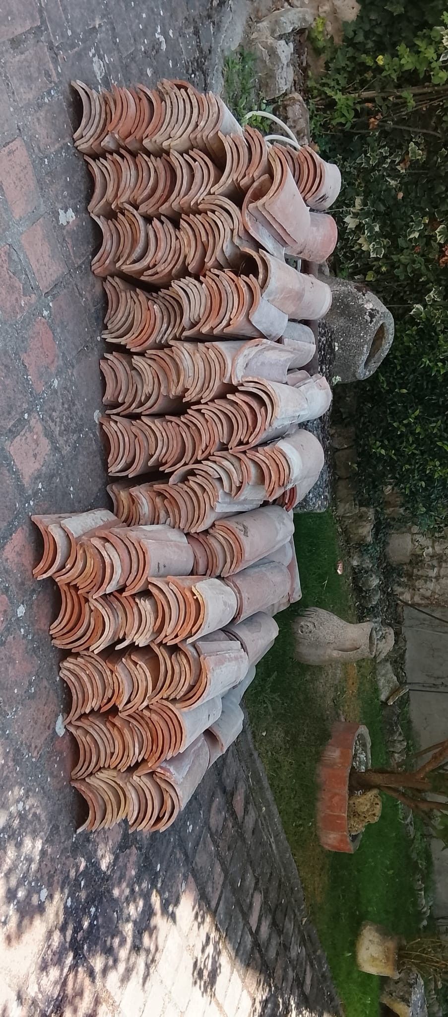 Macetas de barro apiladas secándose al aire libre en un camino de tierra, junto a vegetación y un muro de piedra.