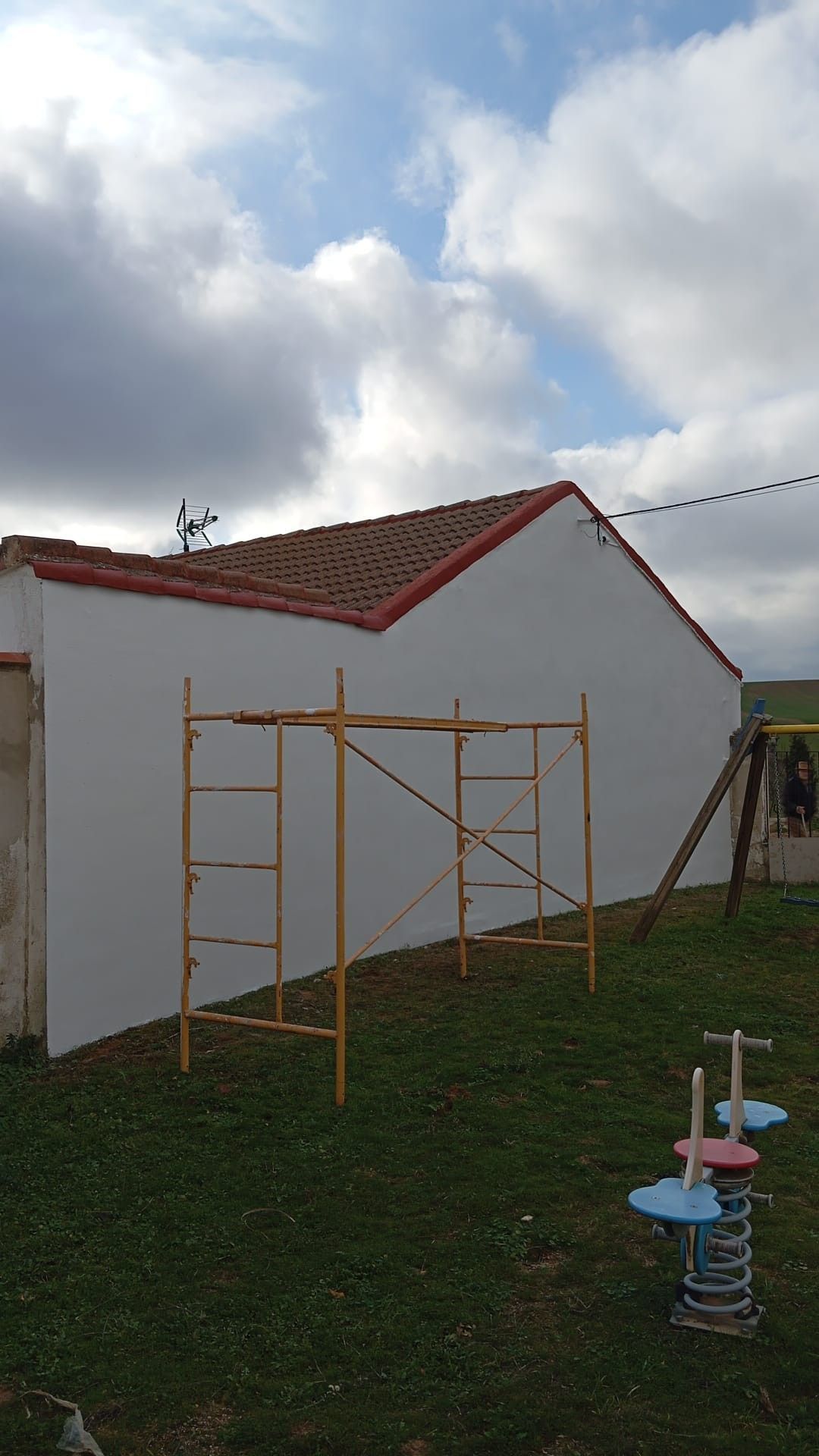 Andamios apoyados contra una pared blanca con tejas rojas, al aire libre en un jardín cubierto de hierba bajo un cielo nublado