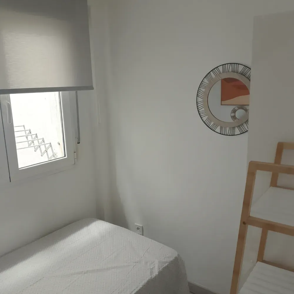 Habitación pequeña de color blanco con cama, ventana, reloj de pared y estantería de madera tipo escalera.