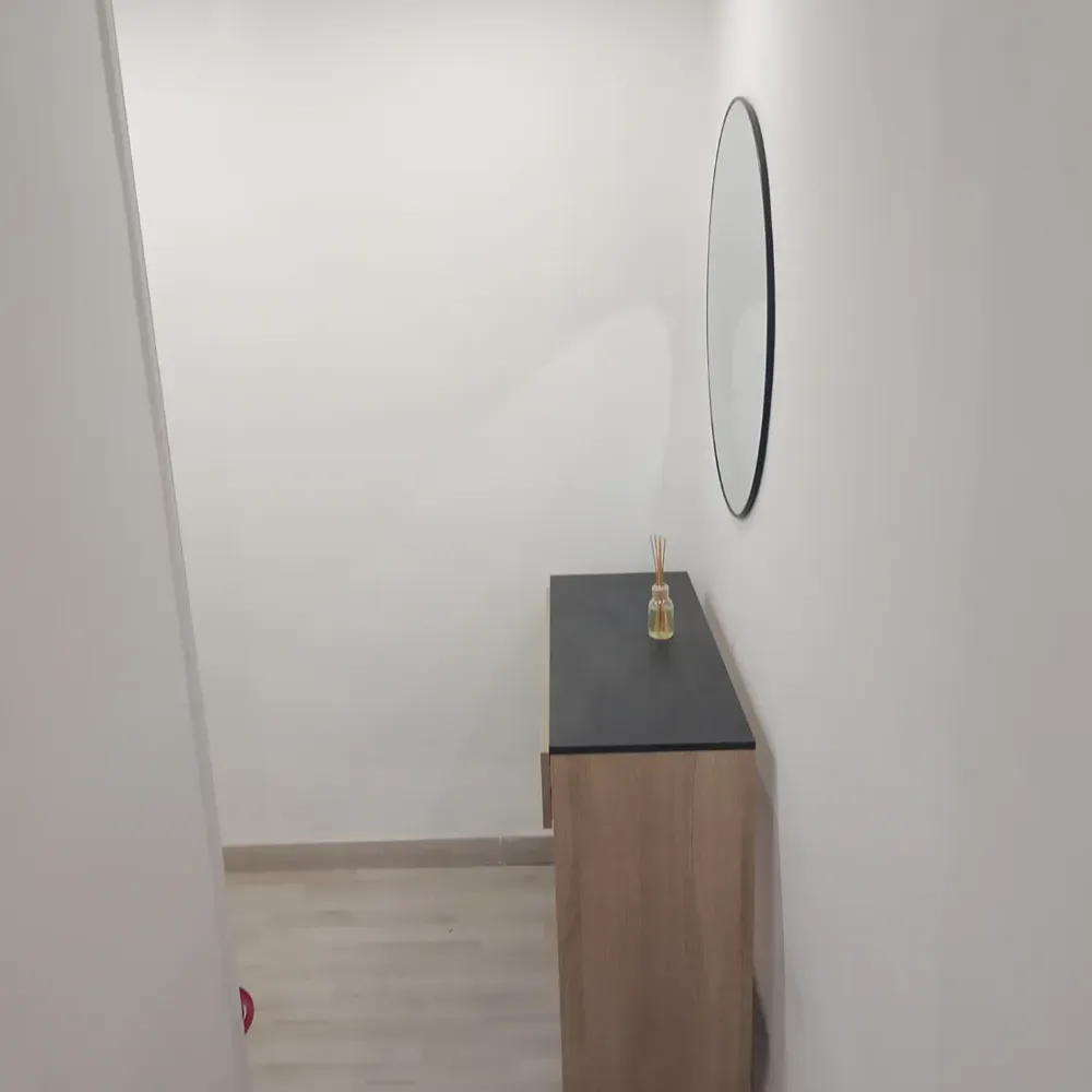 Habitación minimalista con paredes blancas, un pequeño armario de madera y un espejo de pared ovalado negro.
