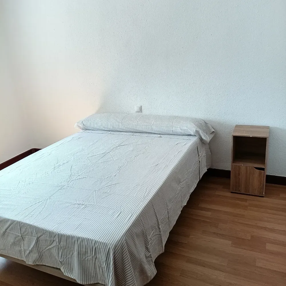 Habitación sencilla con una cama individual, ropa de cama a rayas, suelo de madera y una pequeña mesita de noche.