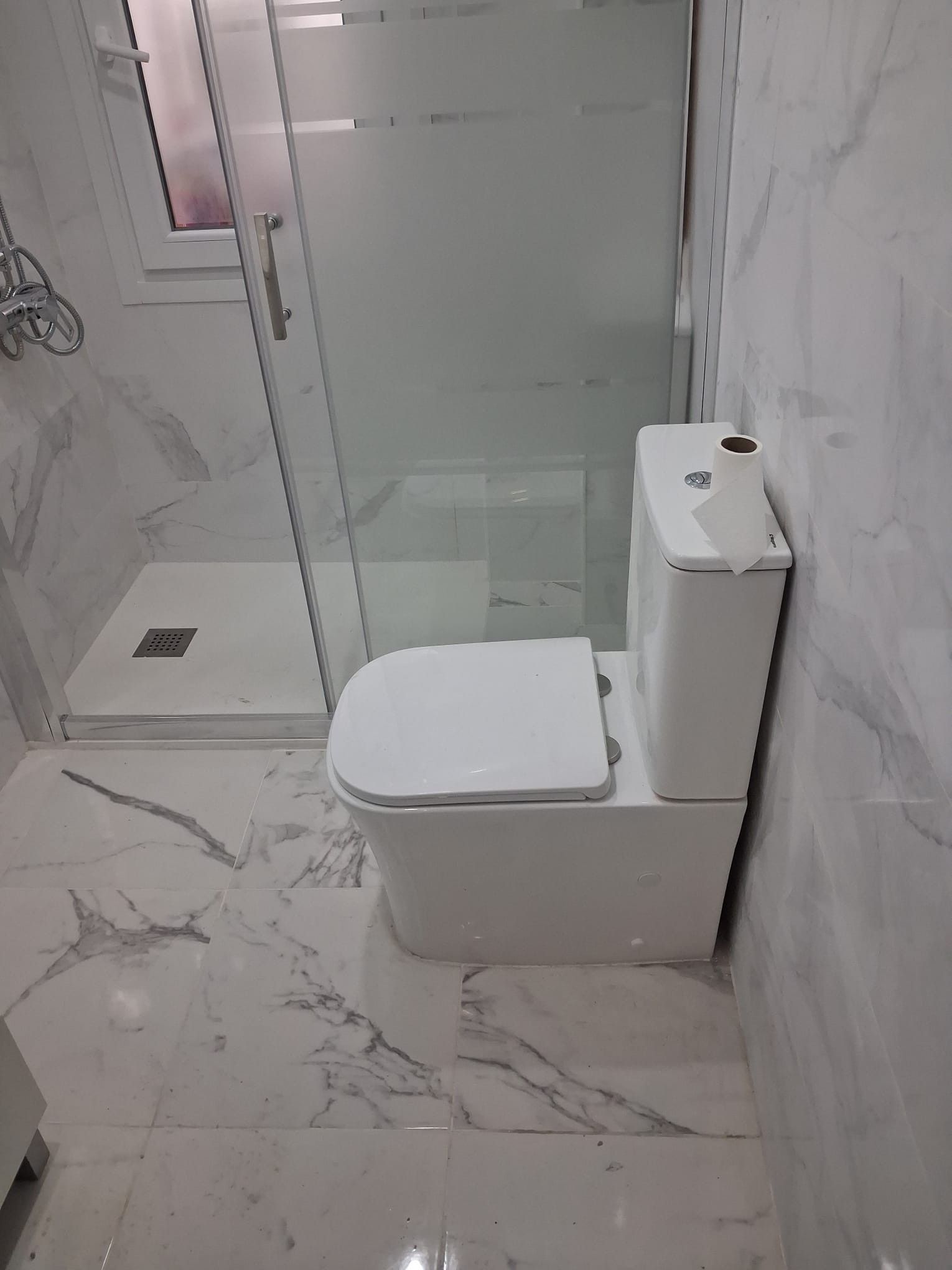 Baño moderno con inodoro blanco y mampara de ducha de cristal sobre azulejos con estampado de mármol.