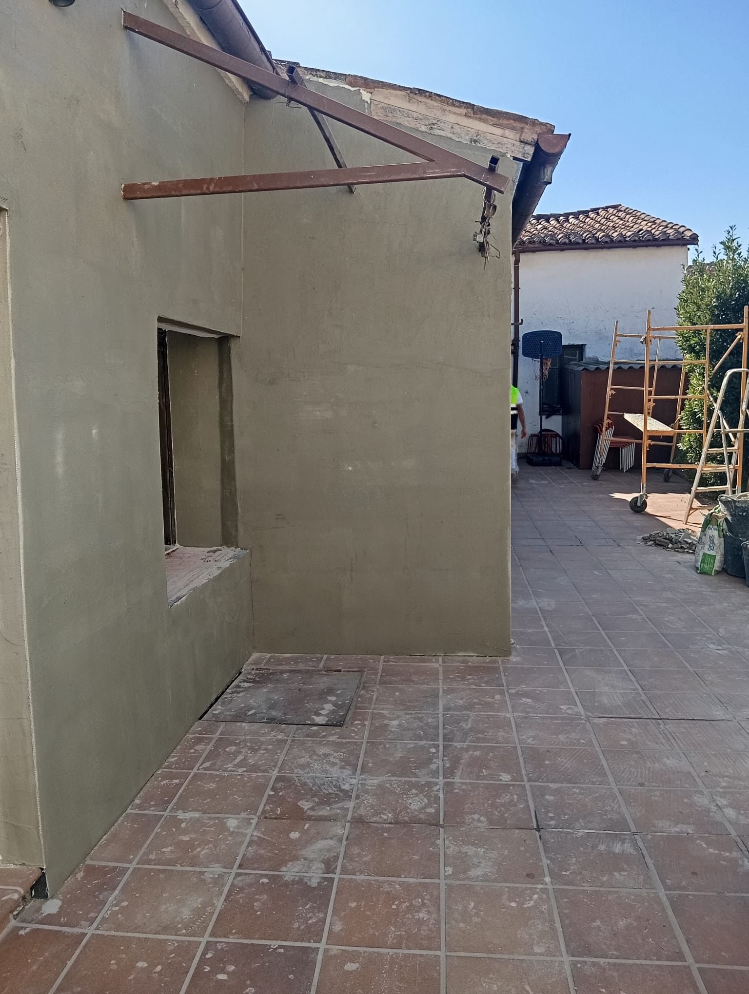 Patio junto a una casa de estuco beige, con suelo de baldosas, una ventana abierta y una zona exterior cubierta al fondo