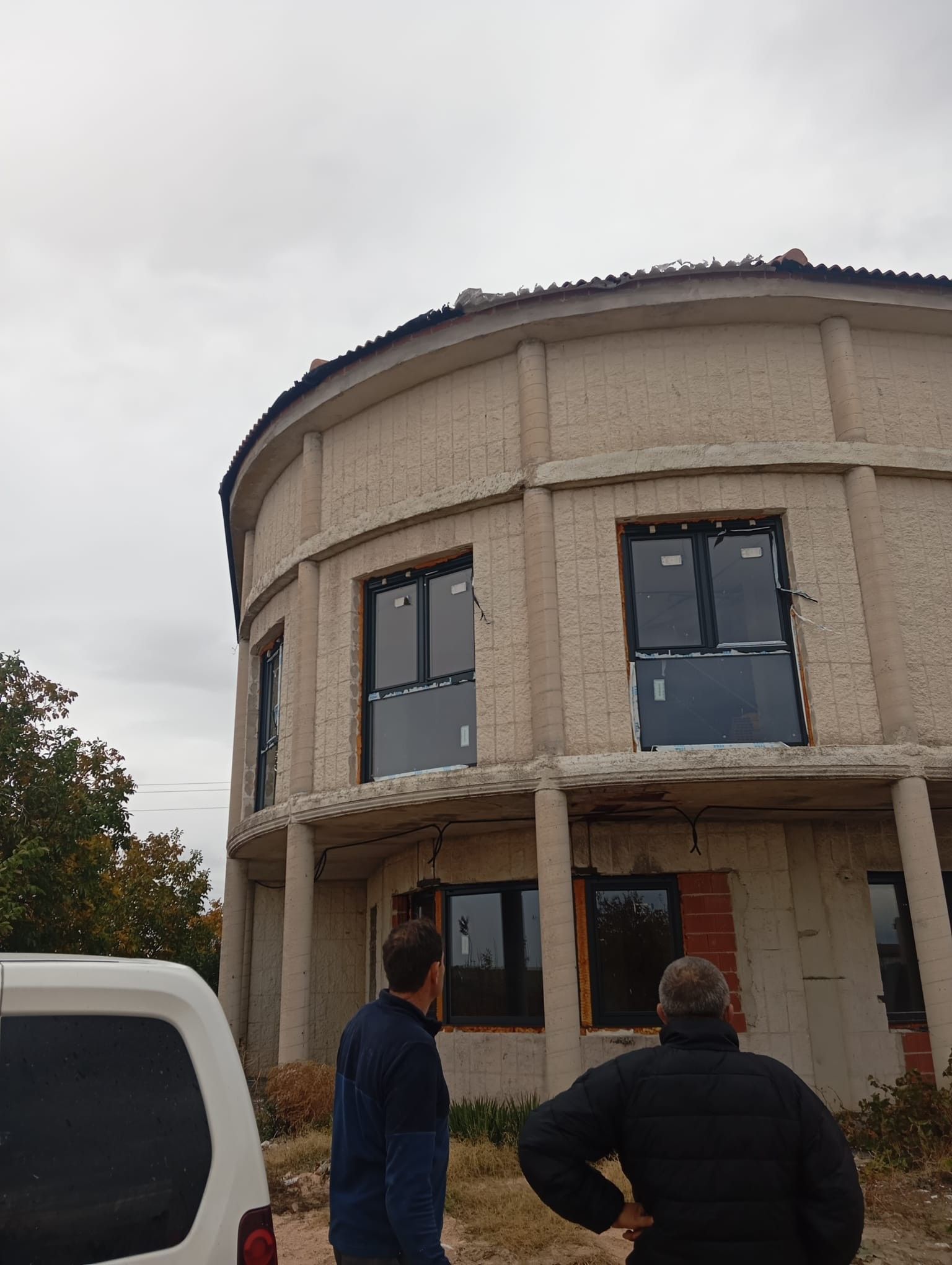 Edificio redondo en construcción, con dos personas de pie delante y una furgoneta blanca a la izquierda.