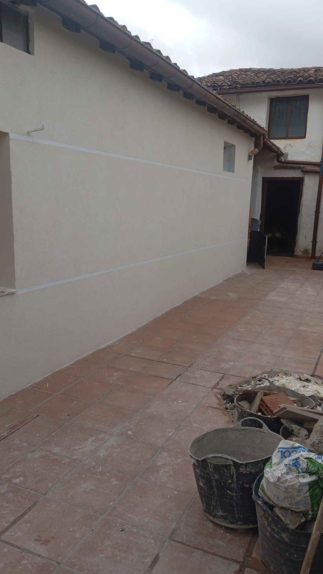 Patio estrecho embaldosado junto a una pared de estuco blanco, con cubos y escombros cerca de una puerta abierta.