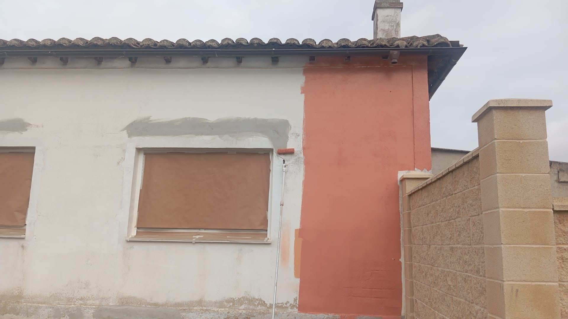 Exterior de una casa con paredes blancas y coral, ventanas con contraventanas y un tejado de tejas bajo un cielo nublado