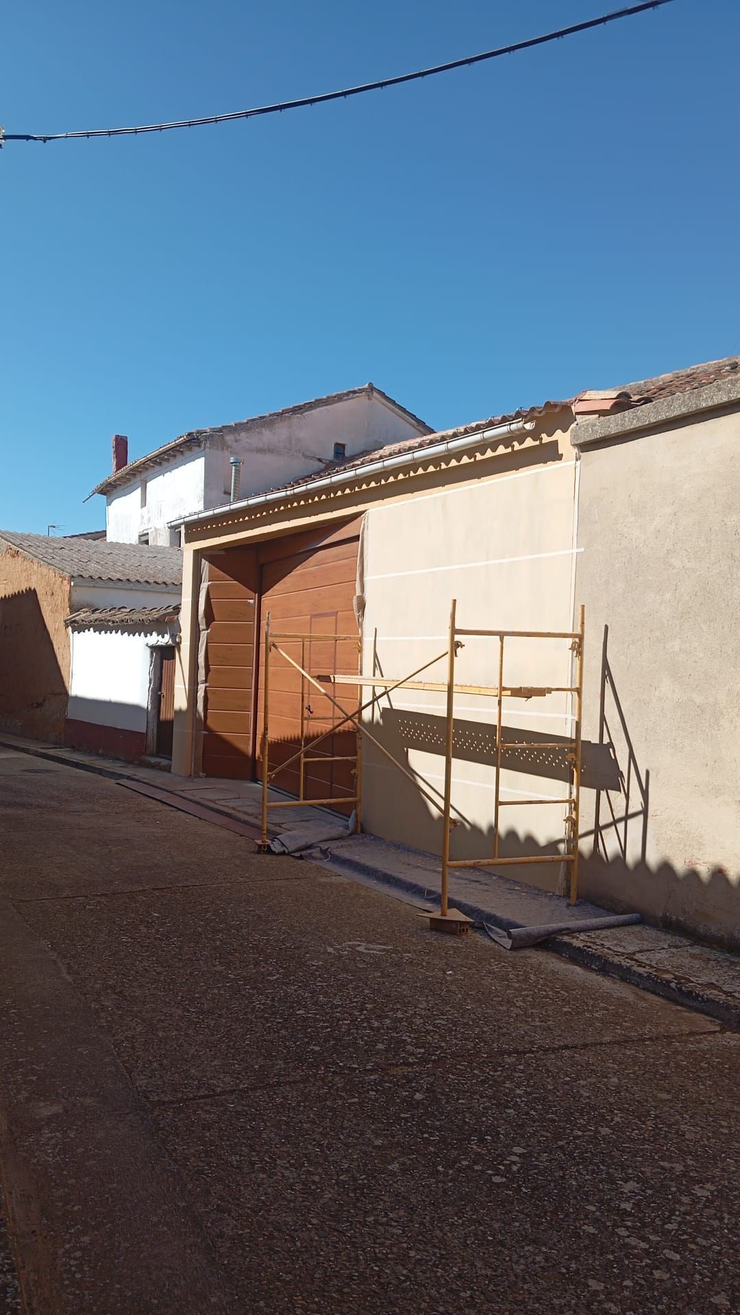Calle soleada con una casa de fachada beige y andamios de madera junto a un camino de grava.