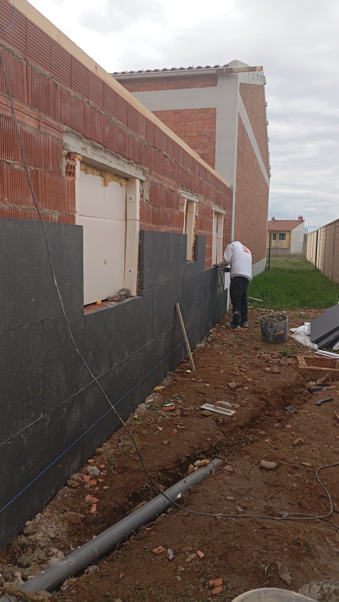 Muro de ladrillo sin terminar junto a una zanja embarrada, con un hombre inspeccionando el lateral de un edificio.