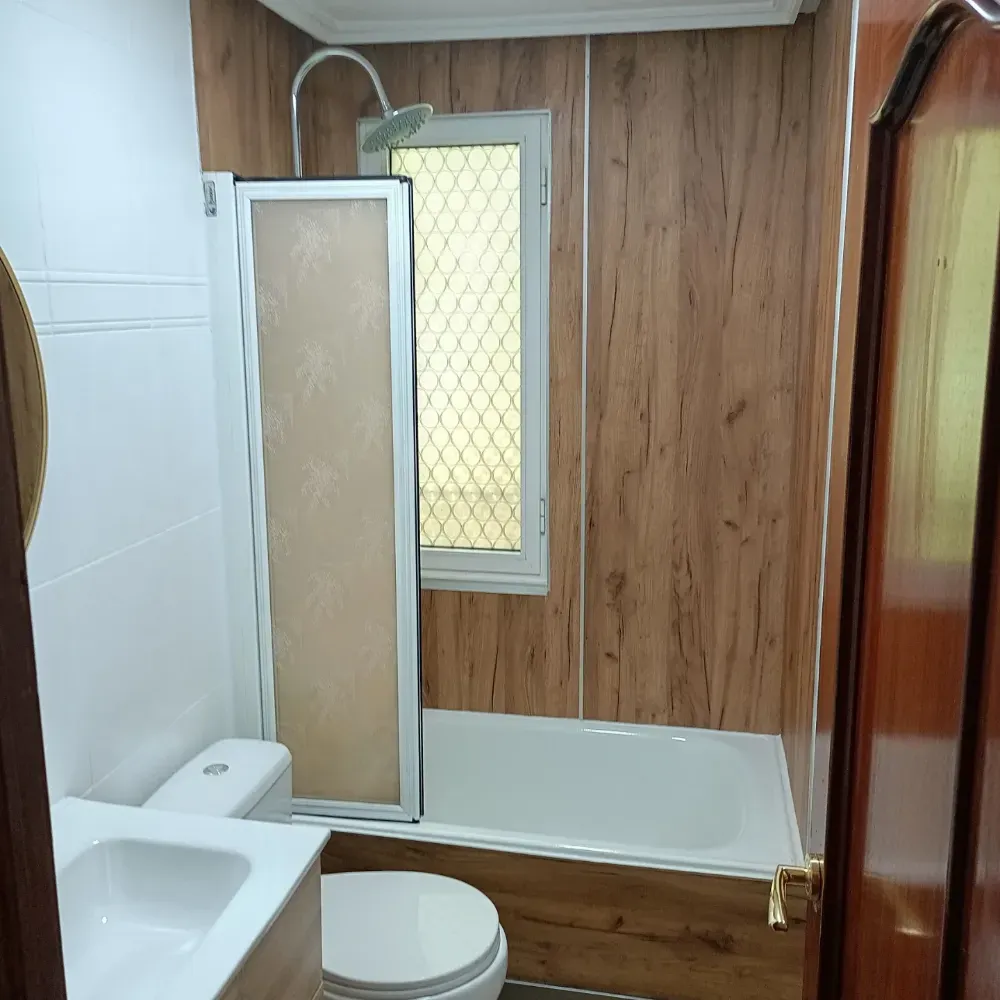 Baño pequeño con inodoro, lavabo, ducha y ventana esmerilada; paredes revestidas de azulejos blancos y paneles de madera.