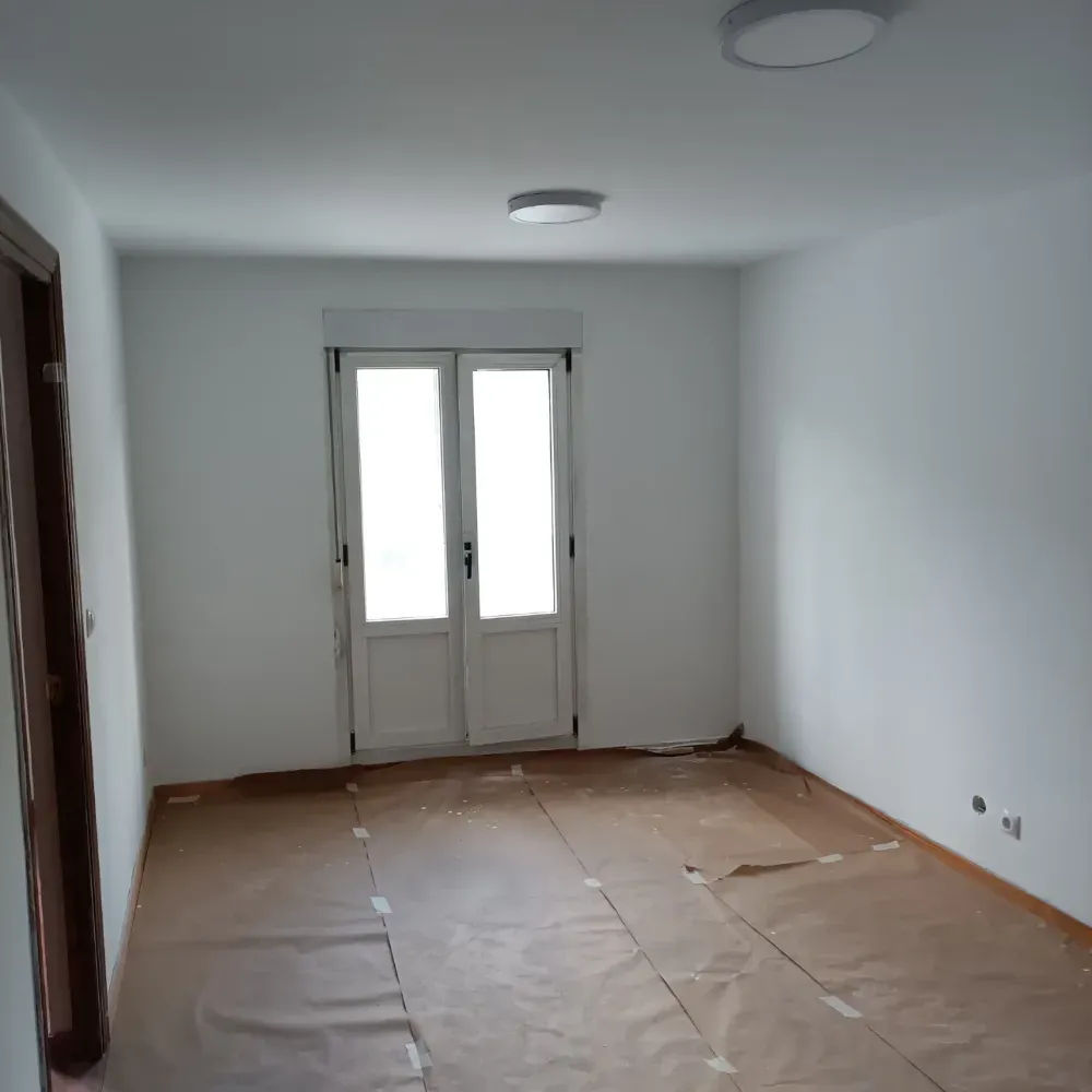Habitación blanca vacía con suelo desnudo, puertas dobles y dos lámparas de techo.