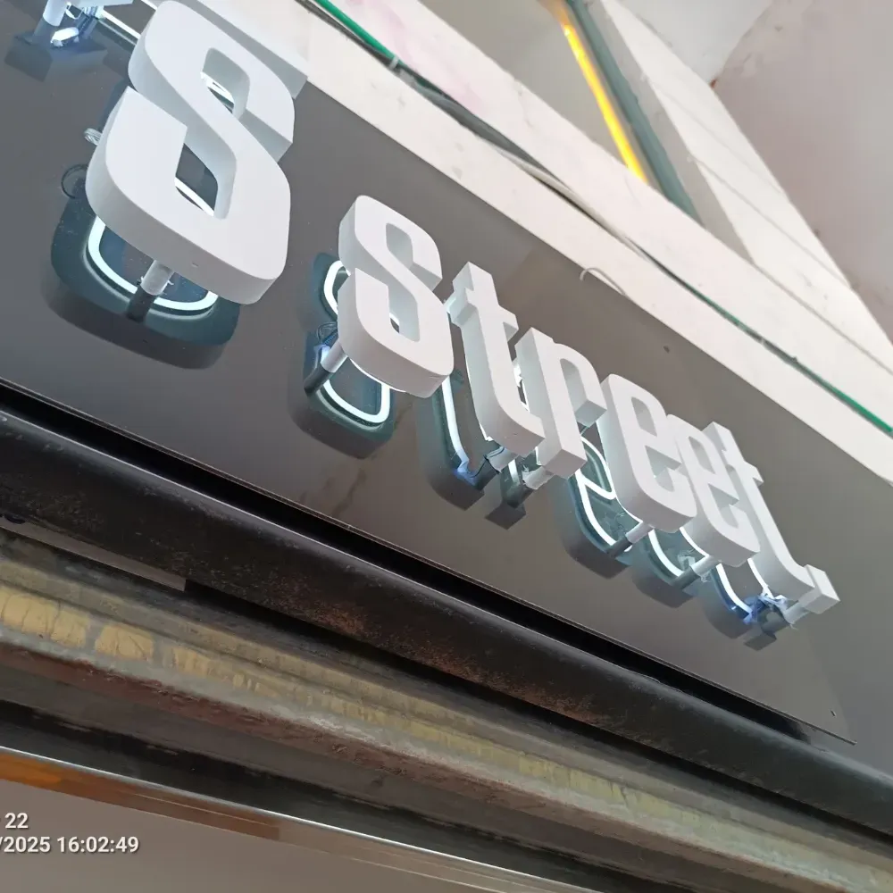 Letrero de la calle 3 sobre un letrero negro en la fachada de una tienda, visto en ángulo, con letras plateadas metálicas y detalles azules.