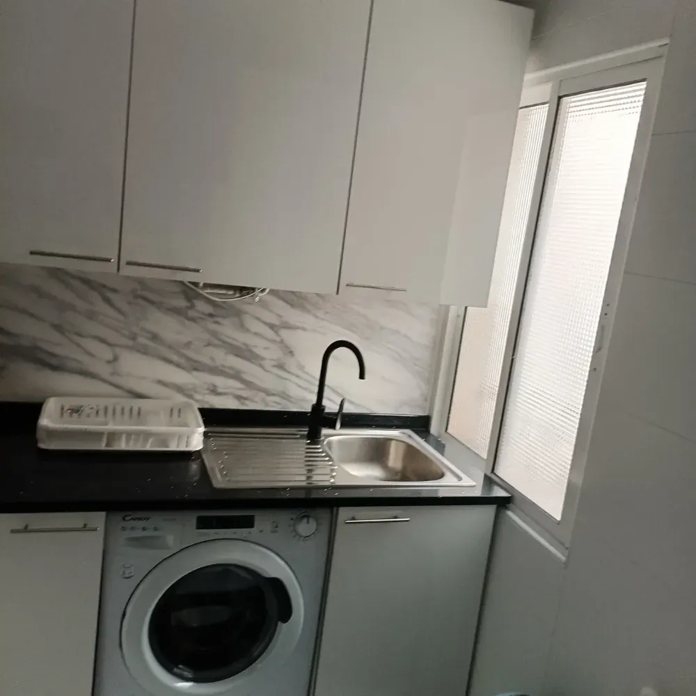 Cocina compacta de color blanco con salpicadero de mármol, encimera negra, fregadero y lavadora bajo la encimera.
