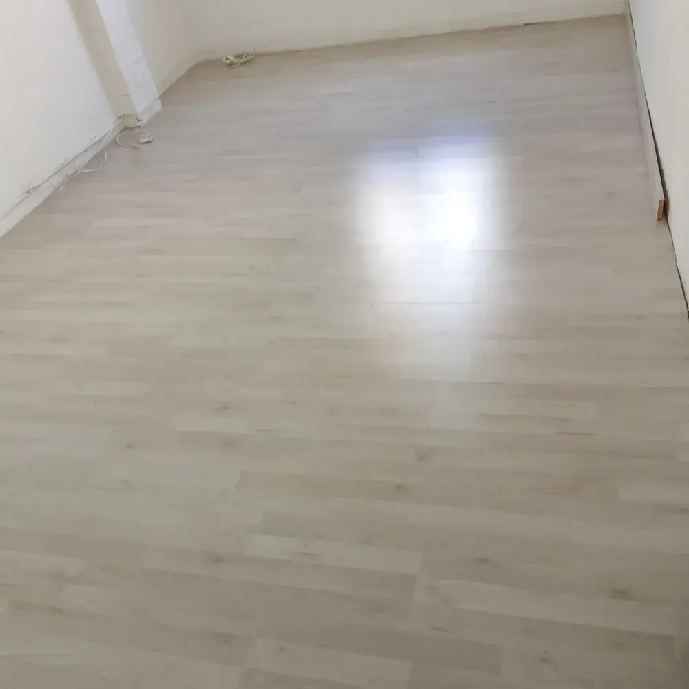 Habitación vacía con suelo de madera clara, paredes blancas y un brillante reflejo en el suelo.