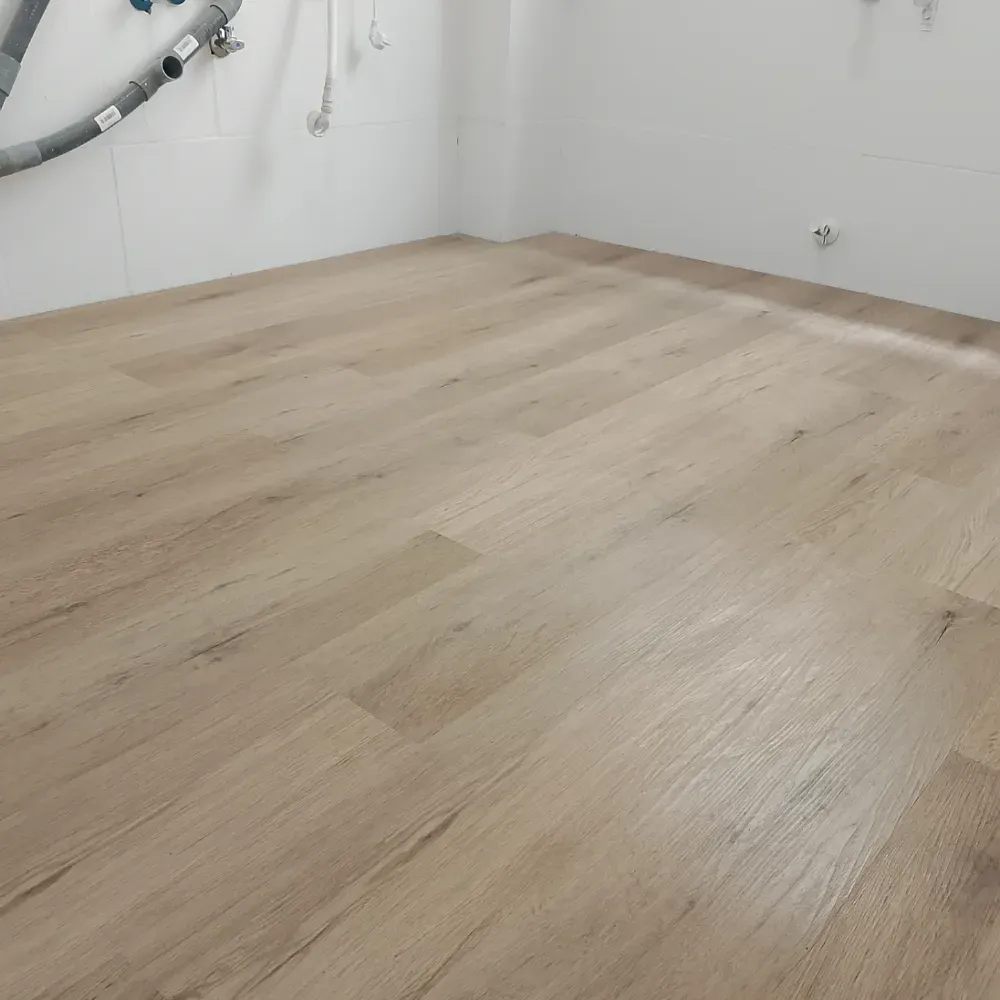 Habitación vacía con suelo de madera clara, paredes blancas y tuberías expuestas en la esquina.