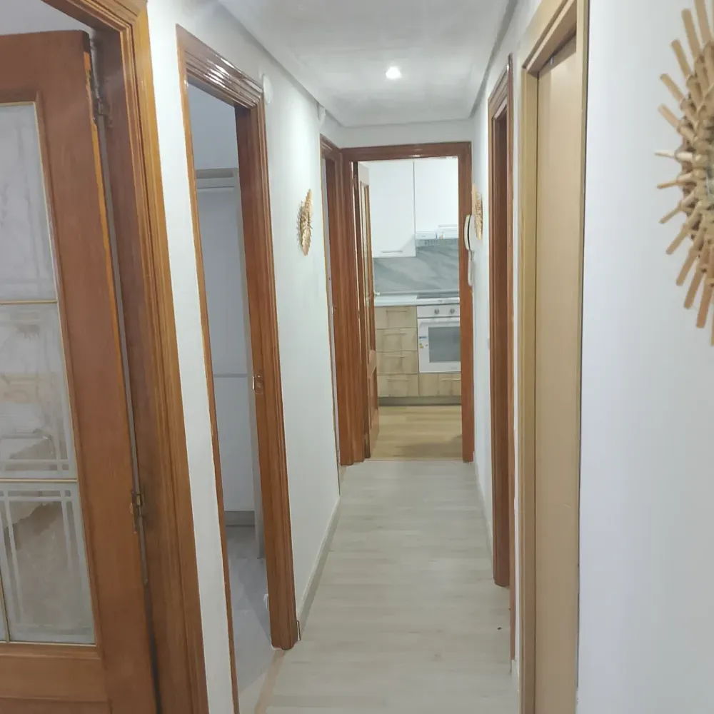 Pasillo estrecho con paredes blancas, puertas de madera y una vista parcial de la cocina al final.