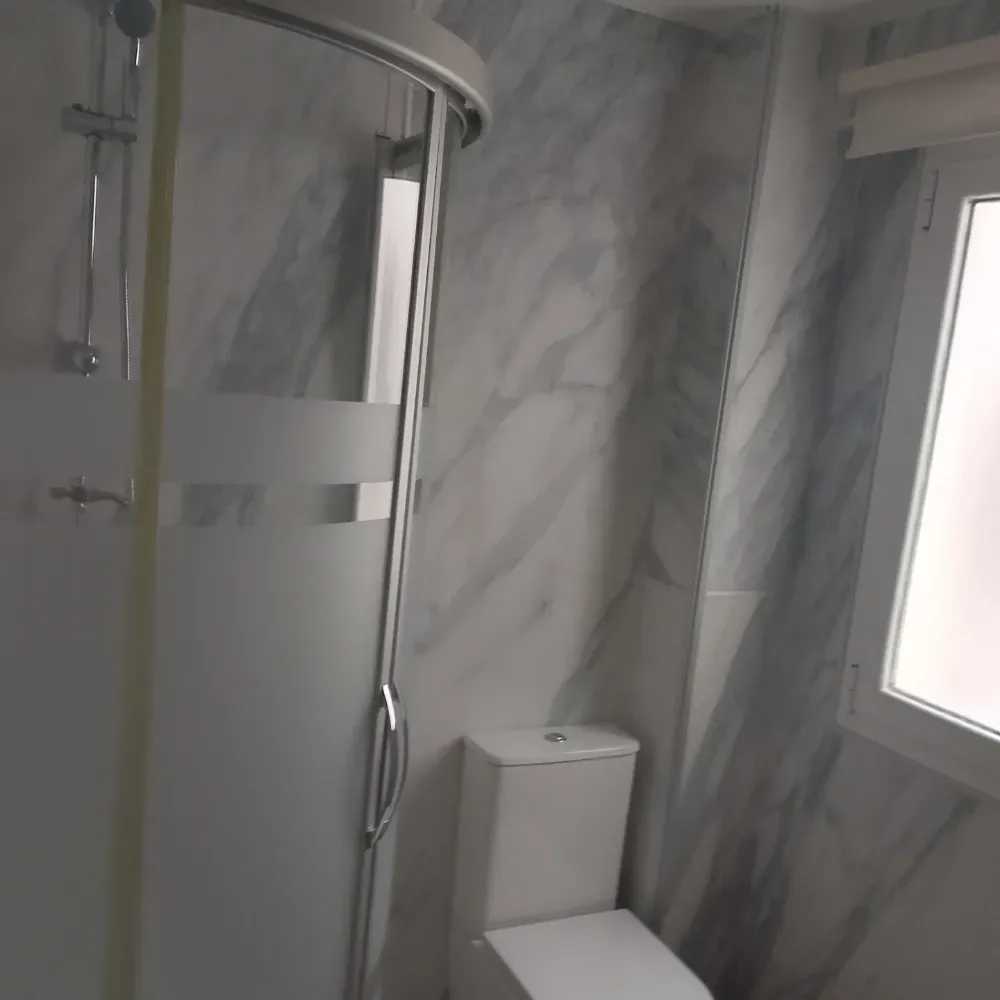 Baño pequeño con cabina de ducha de cristal, paredes con diseño de mármol, inodoro y ventana esmerilada.