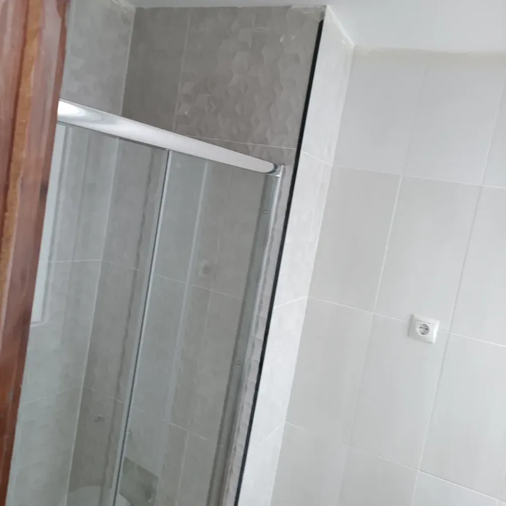 Baño con cabina de ducha de cristal, paredes de azulejos grises y pared de azulejos blancos con toma de corriente.