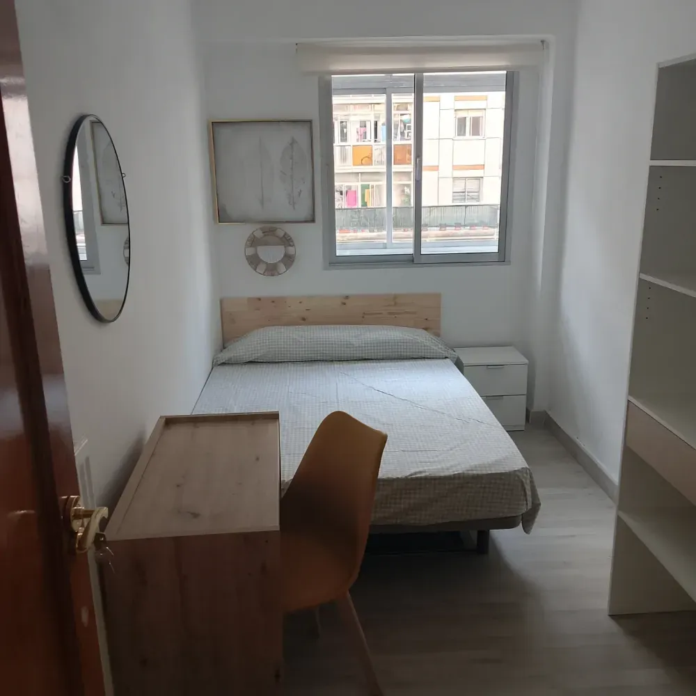 Habitación pequeña y luminosa con cama, escritorio, silla, espejo, ventana y estanterías blancas.
