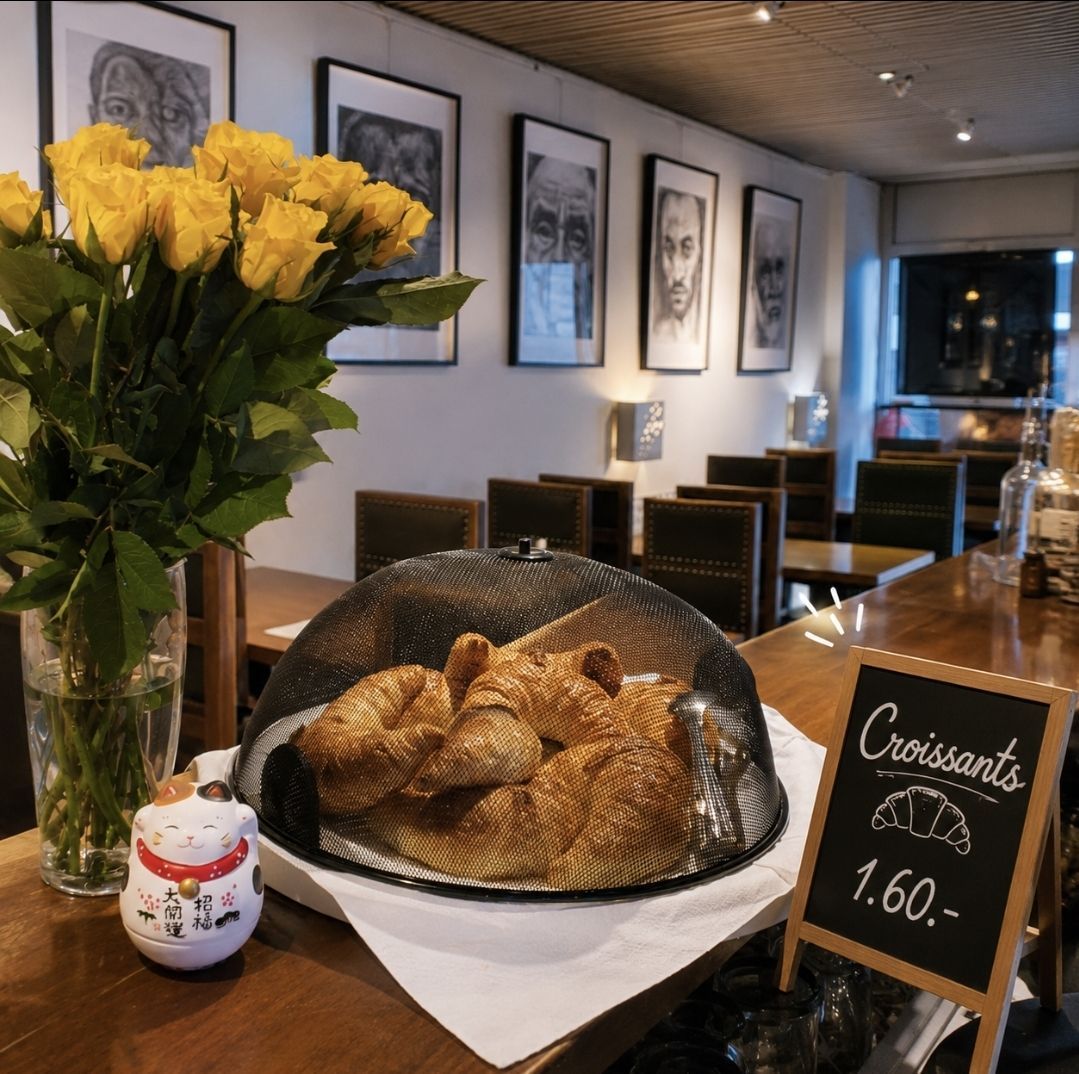 Comptoir de boulangerie avec des croissants sous un dôme, des fleurs jaunes et une pancarte indiquant « Croissants 1,60 ».