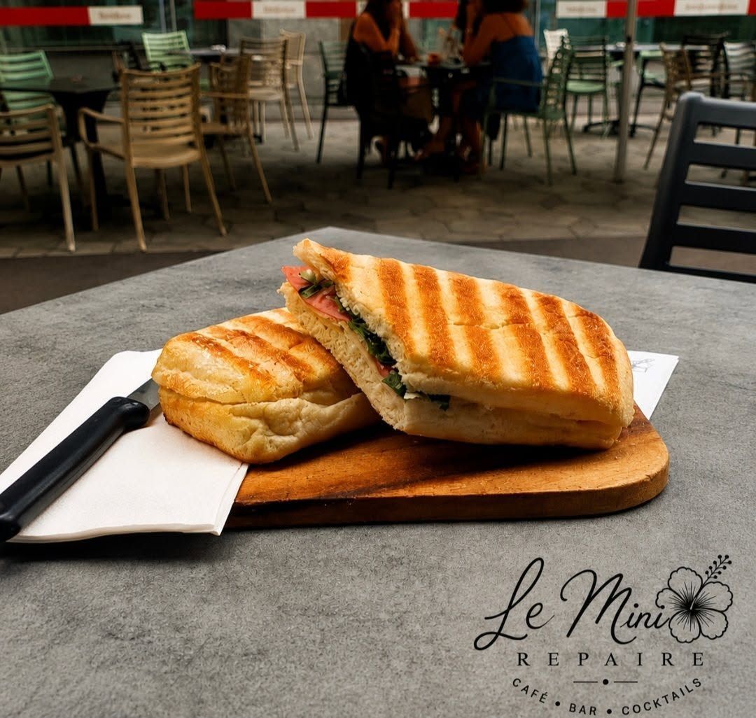 Panini grillé sur une planche en bois, servi à la terrasse d'un café, avec des serviettes et un couteau.