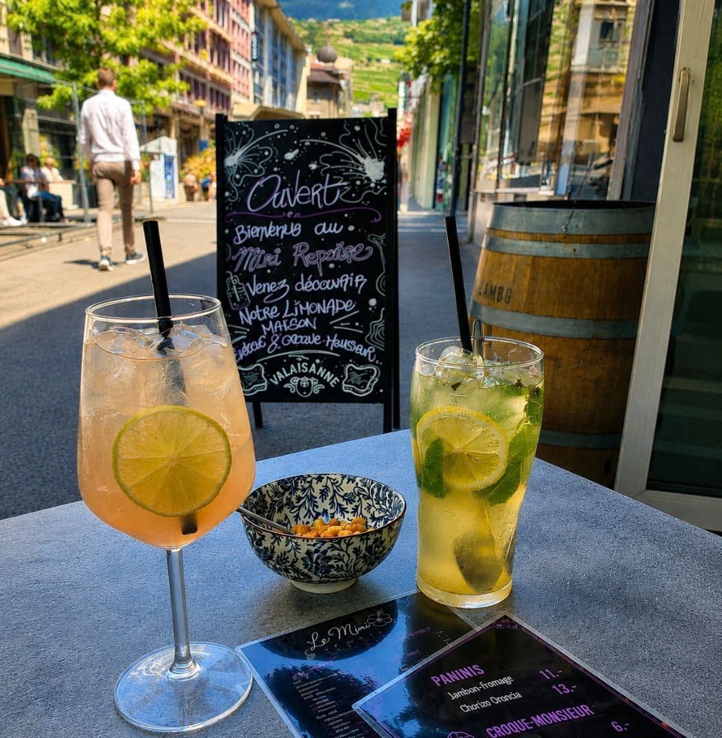 Deux cocktails colorés sur une table de café, à côté d'un menu écrit à la craie sur un tableau noir, en terrasse.