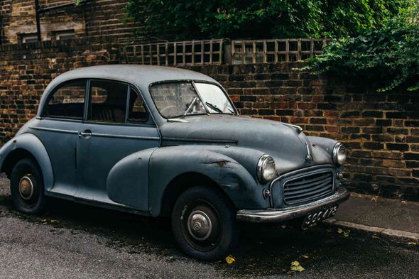 Un coche antiguo, de color azul y desgastado, aparcado en una calle junto a una pared de ladrillos.