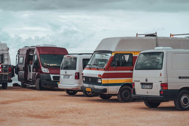 Furgonetas estacionadas en una playa de arena. Varios colores: blanco, rojo, amarillo. Cielo nublado.