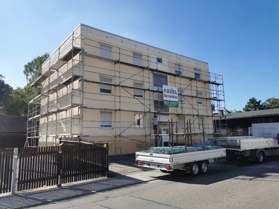 Gebäude im Umbau mit Gerüst, beige Fassade, davor Anhänger mit Vorräten.