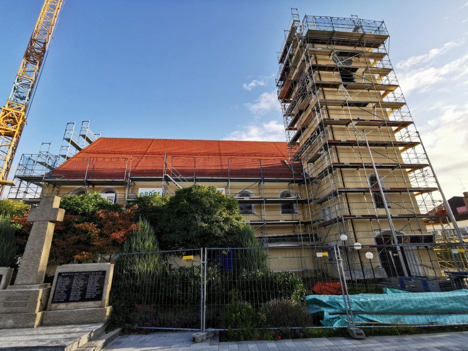 Kirche wird renoviert; Gerüst umgibt den Turm. Rotes Dach, beige Wände, Kreuz und Kriegerdenkmal.