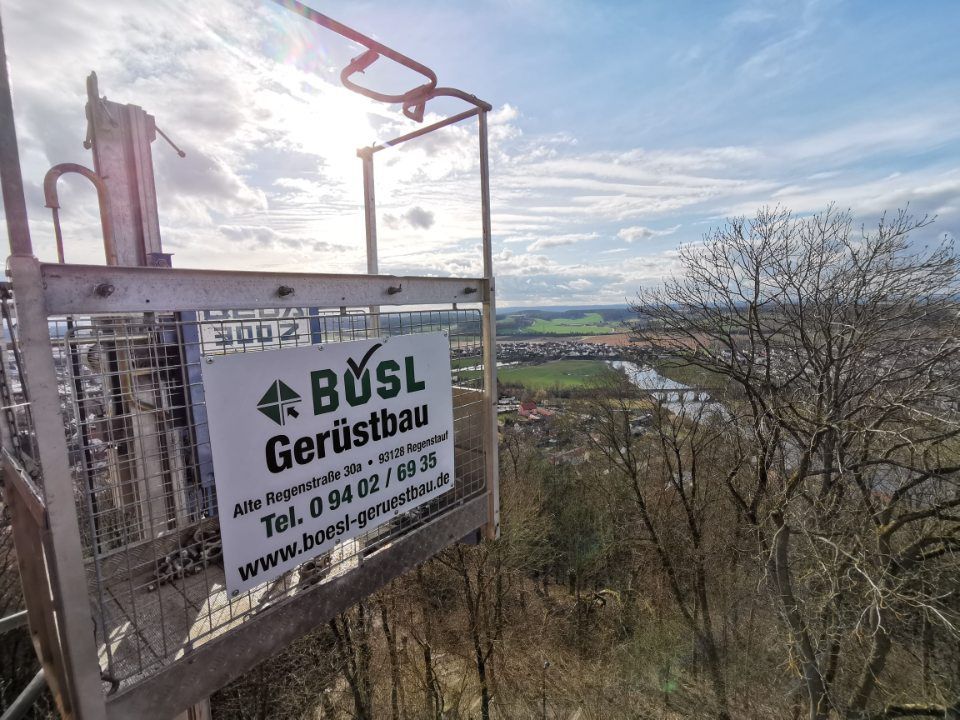 Gerüstplattform mit Schild „BÜSL Gerüstbau“ mit Blick auf eine entfernte Stadt und einen Fluss an einem sonnigen Tag.