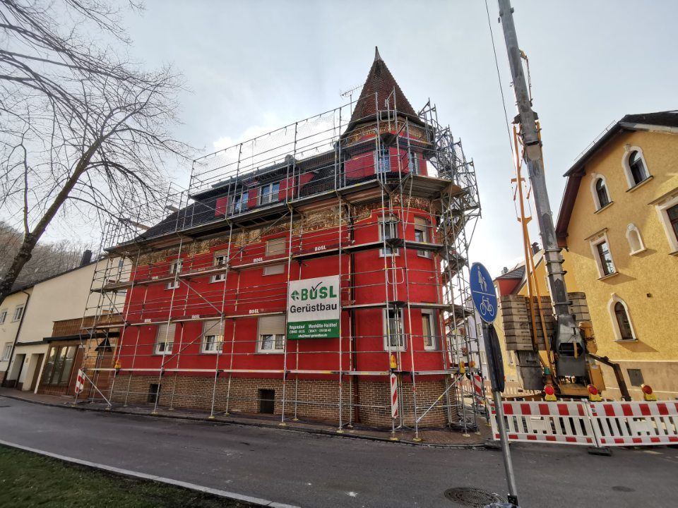 Gebäude im Umbau; rote Fassade, Gerüst, Kran. Schild der Baufirma an der Seite.