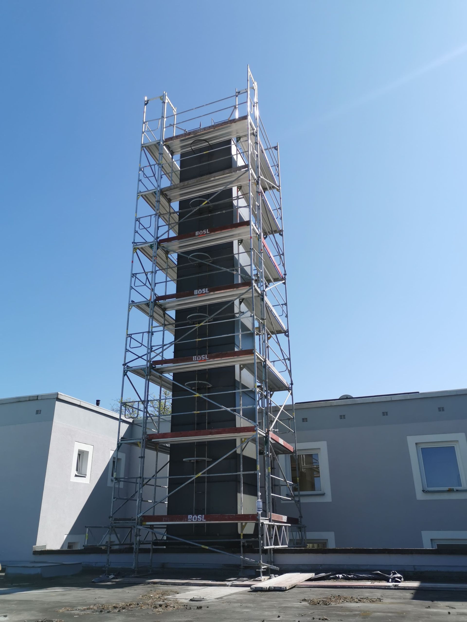 Ein Gerüst umgibt ein hohes, dunkles Bauwerk neben einem niedrigen Gebäude unter blauem Himmel.