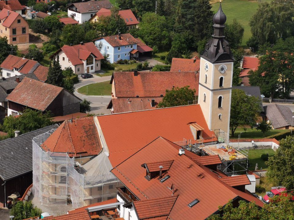 Eine Kirche mit einem hohen Glockenturm in einem europäischen Dorf, umgeben von Häusern mit roten Ziegeldächern.