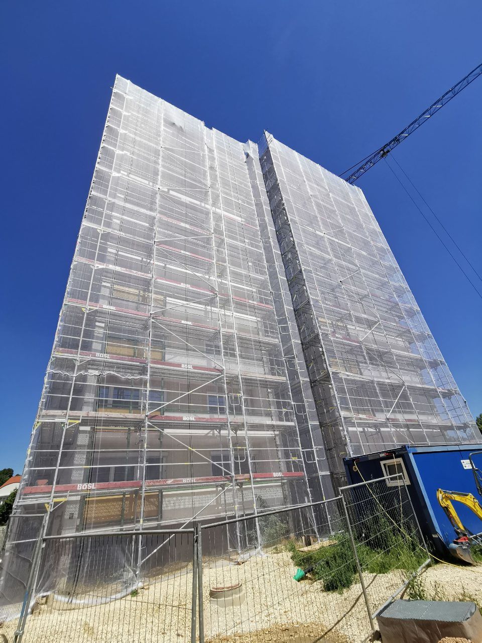 Ein Gerüst bedeckt ein hohes Gebäude im Bau, blauen Himmel, Kran und Baumaschinen.