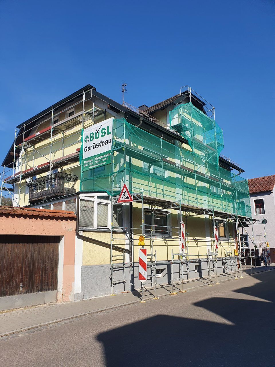 Gebäude im Umbau mit Gerüst und Sicherheitsnetz an einer Straße unter blauem Himmel.