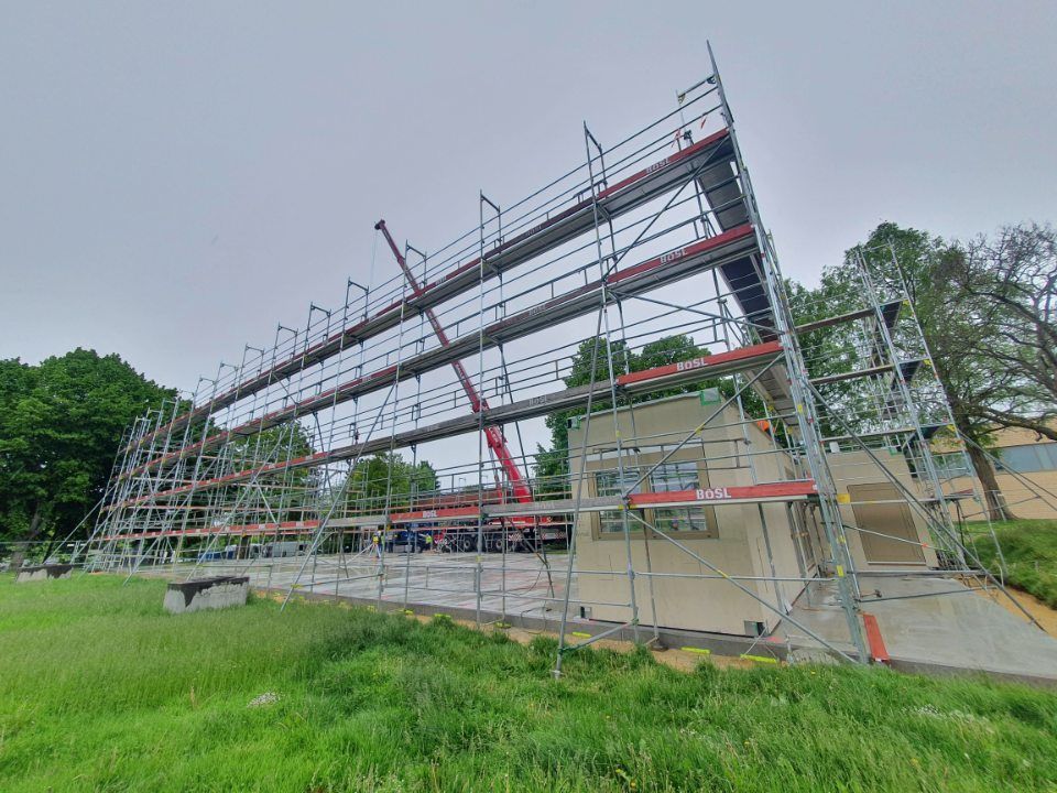 An der Gebäudeaußenseite ist ein Gerüst errichtet, im Hintergrund ein roter Kran, im Vordergrund grünes Gras, bewölkter Tag.