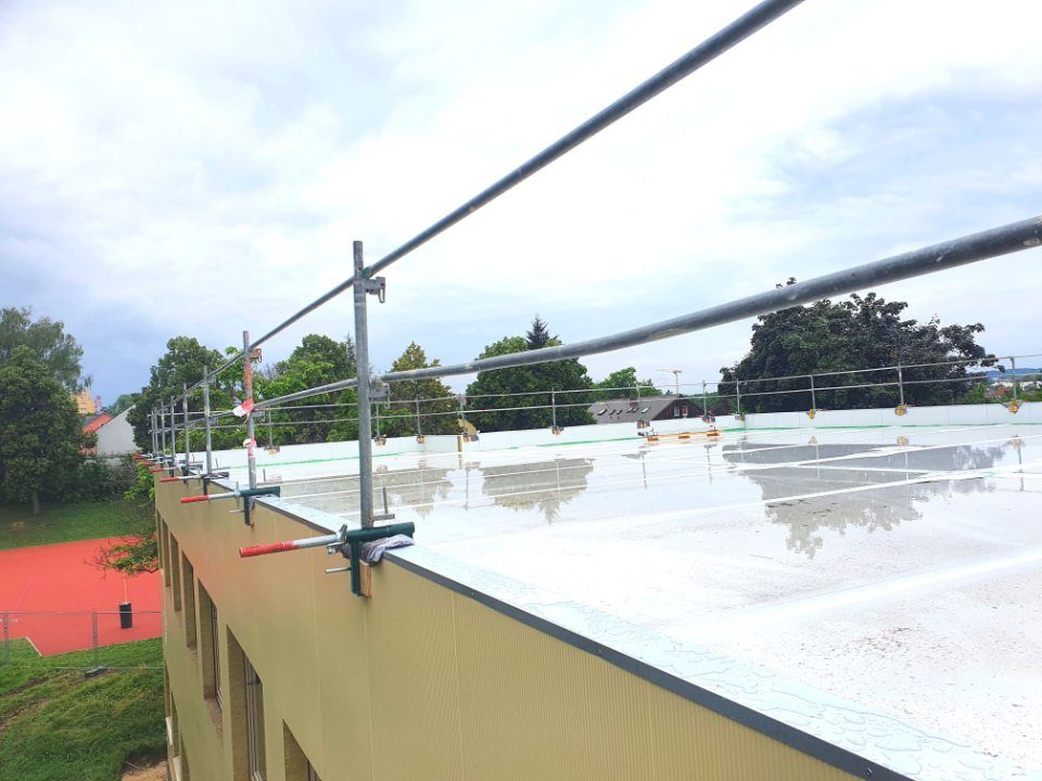 Dach mit silbernem Sicherheitsgeländersystem. Grünes Gras und Bäume im Hintergrund.