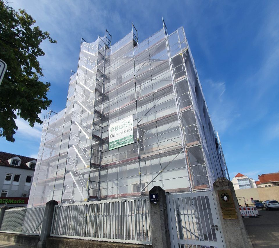 Gebäude im Umbau, mit Gerüsten und Netzen bedeckt. Weißes und graues Gebäude unter blauem Himmel.
