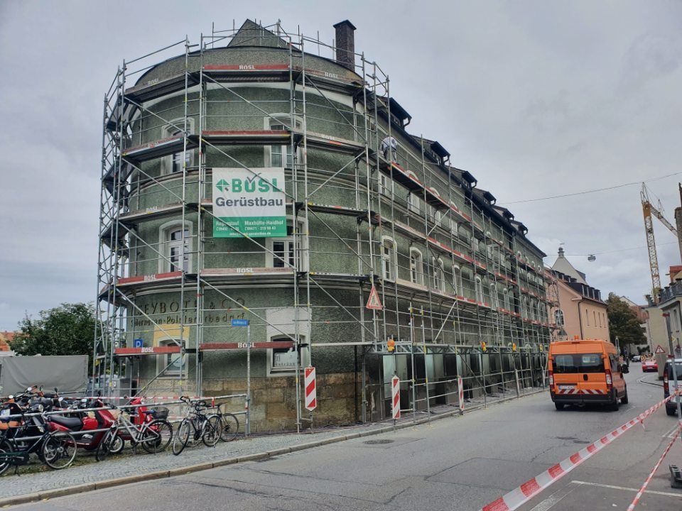 Gebäude wird renoviert, ist mit einem Gerüst bedeckt; orangefarbener Nutzlastwagen parkt auf der Straße.