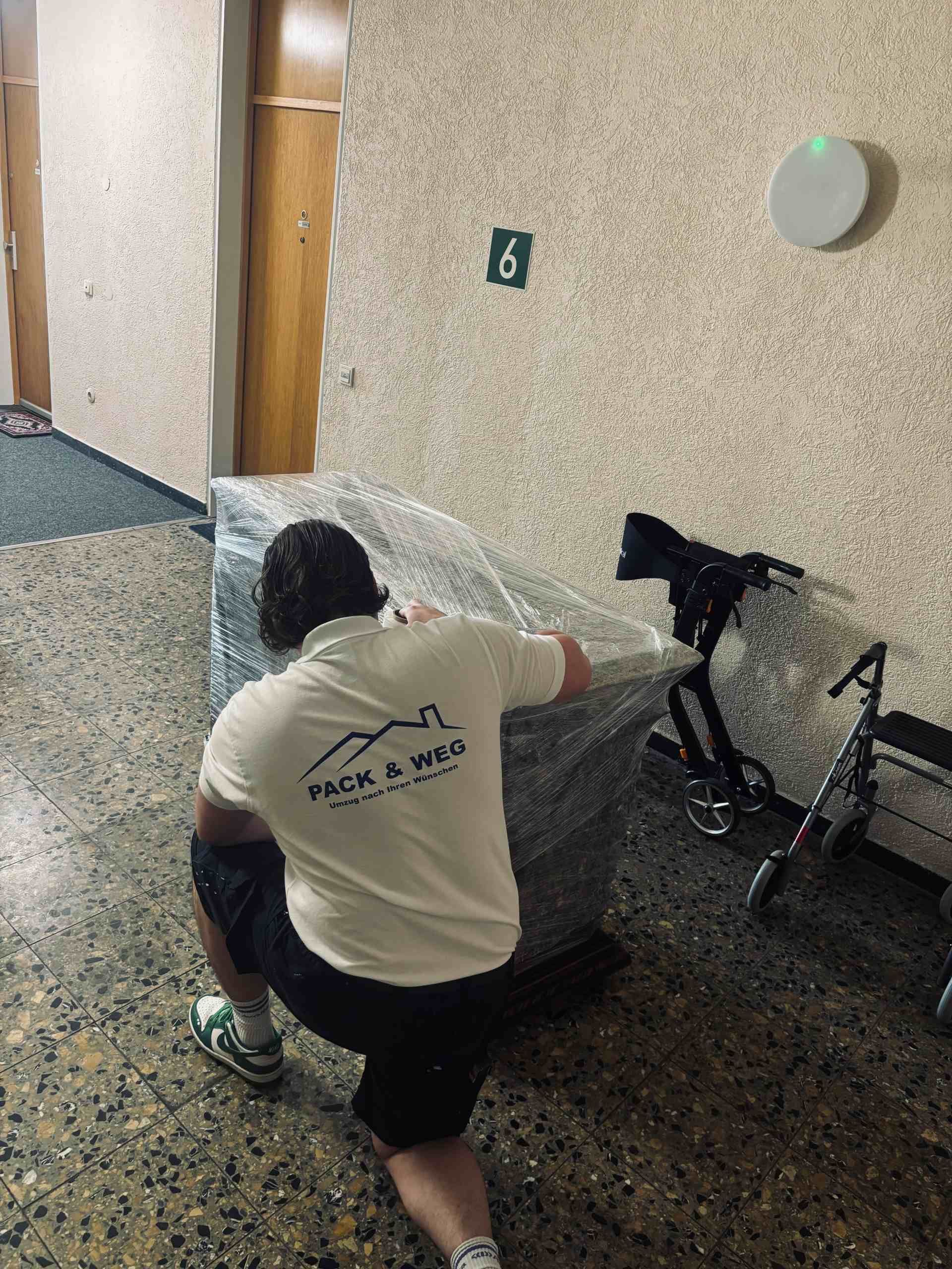 Eine Person kniet und sichert einen großen, verpackten Gegenstand in einem Gebäudeflur. „Pack & Weg“-Logo auf dem Hemd.