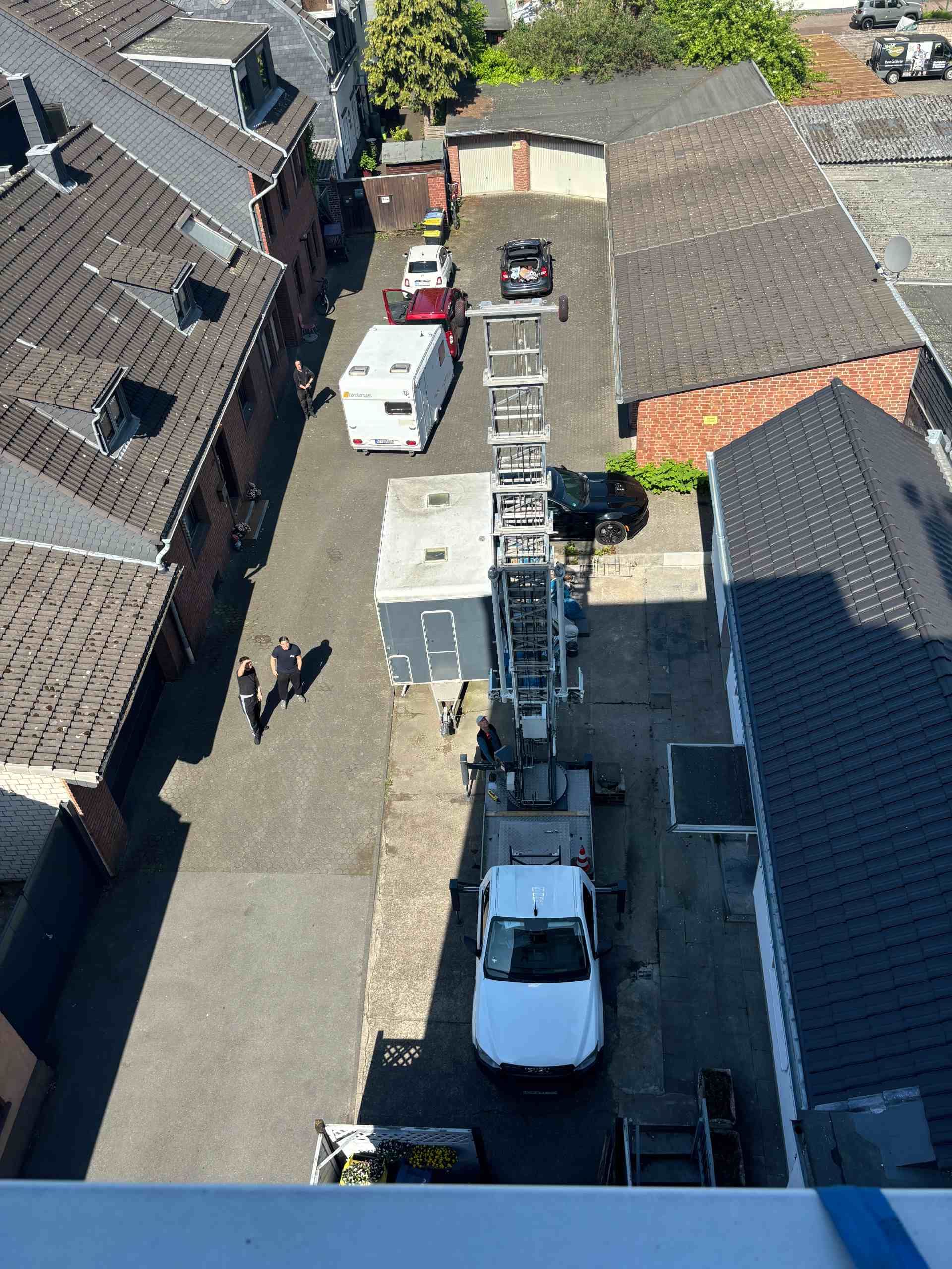 Schmale Gasse mit Gebäuden, Fahrzeugen und einem hohen Drehleiterwagen. Menschen sind anwesend.