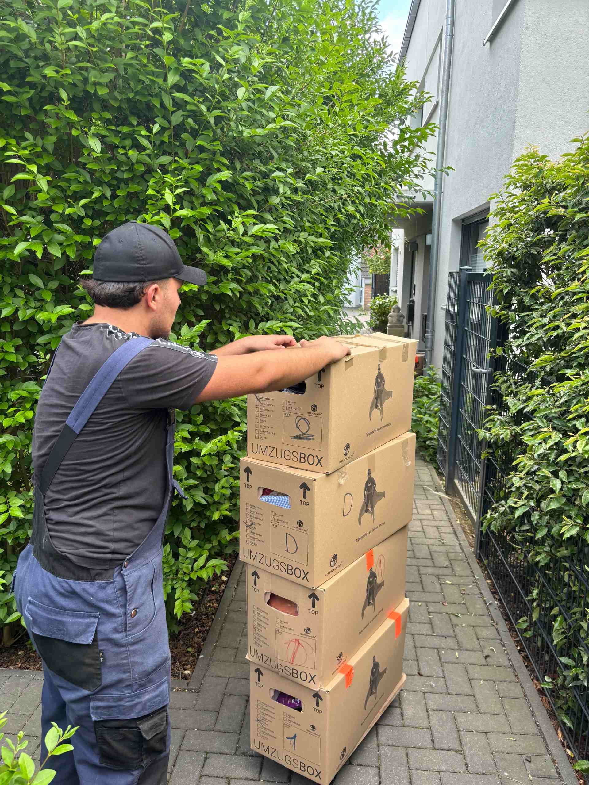 Mann trägt einen Stapel Umzugskartons einen Ziegelweg entlang, in der Nähe eines Hauses und von Büschen.