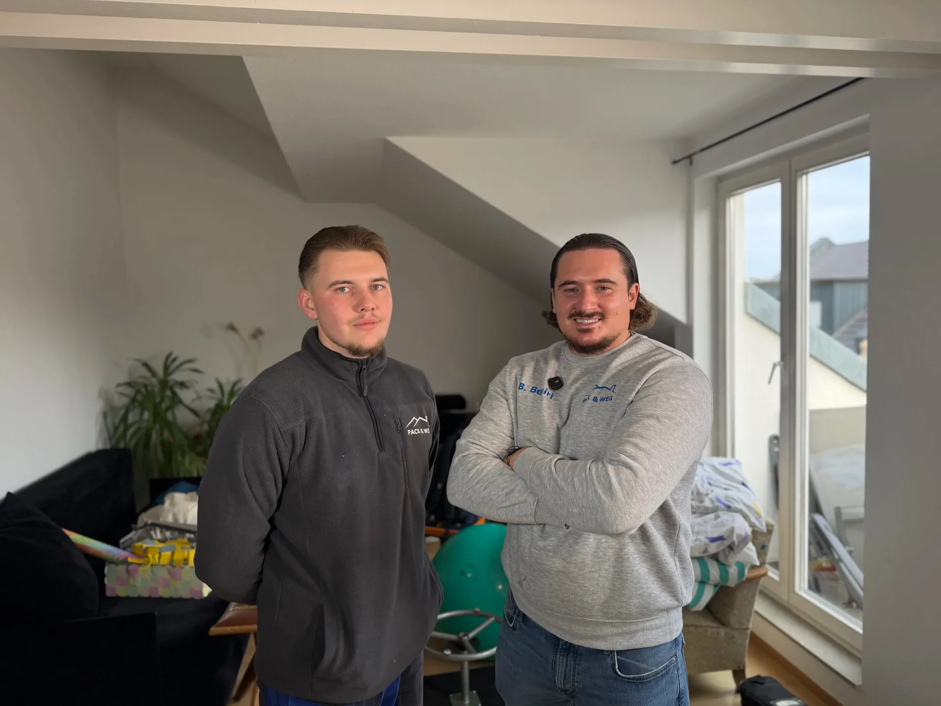 Zwei Männer stehen in einem Innenraum, einer mit verschränkten Armen.