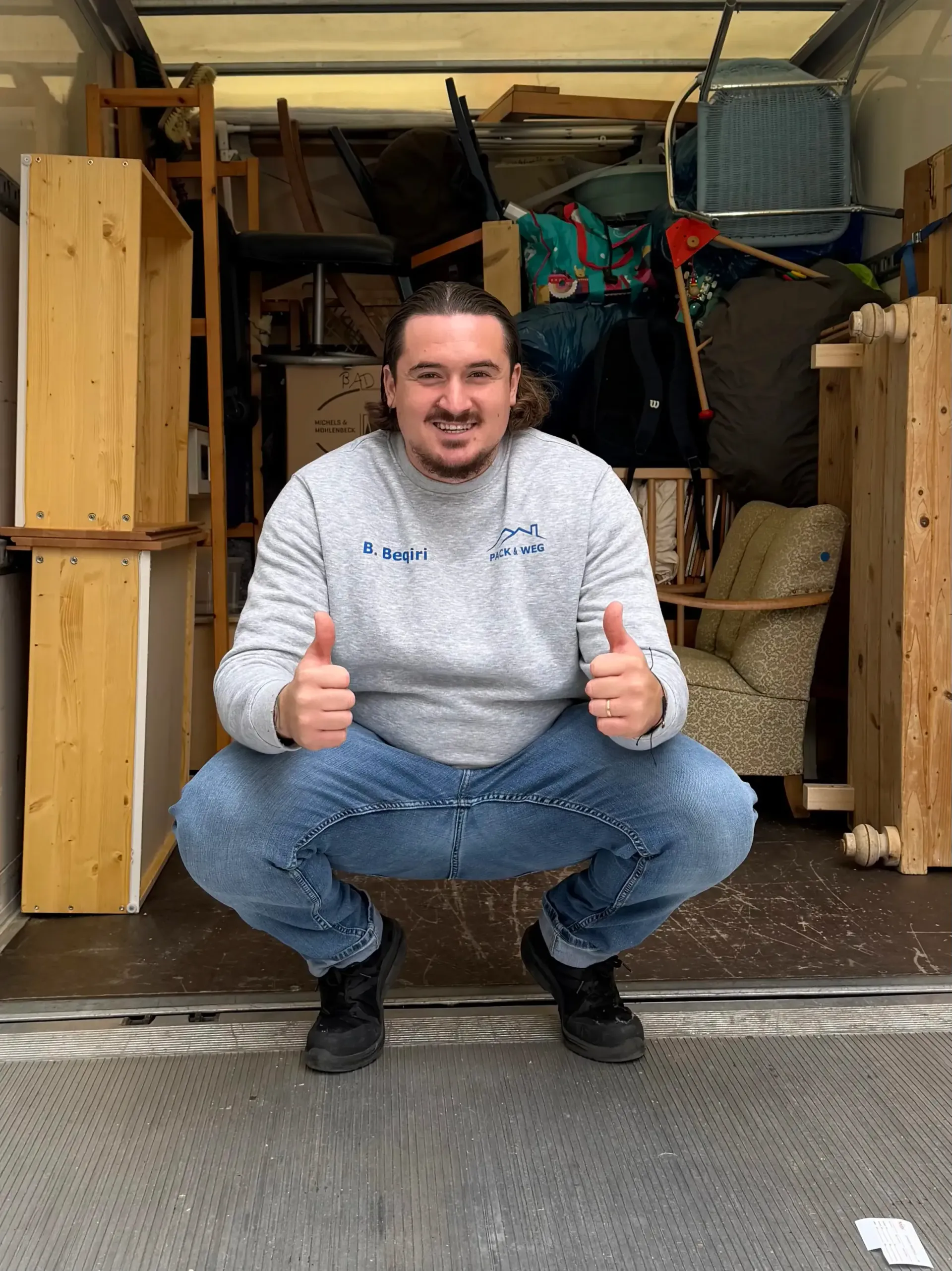 Ein Mann in grauem Pullover und Jeans hockt in einem vollbesetzten Umzugswagen und zeigt zwei Daumen nach oben.