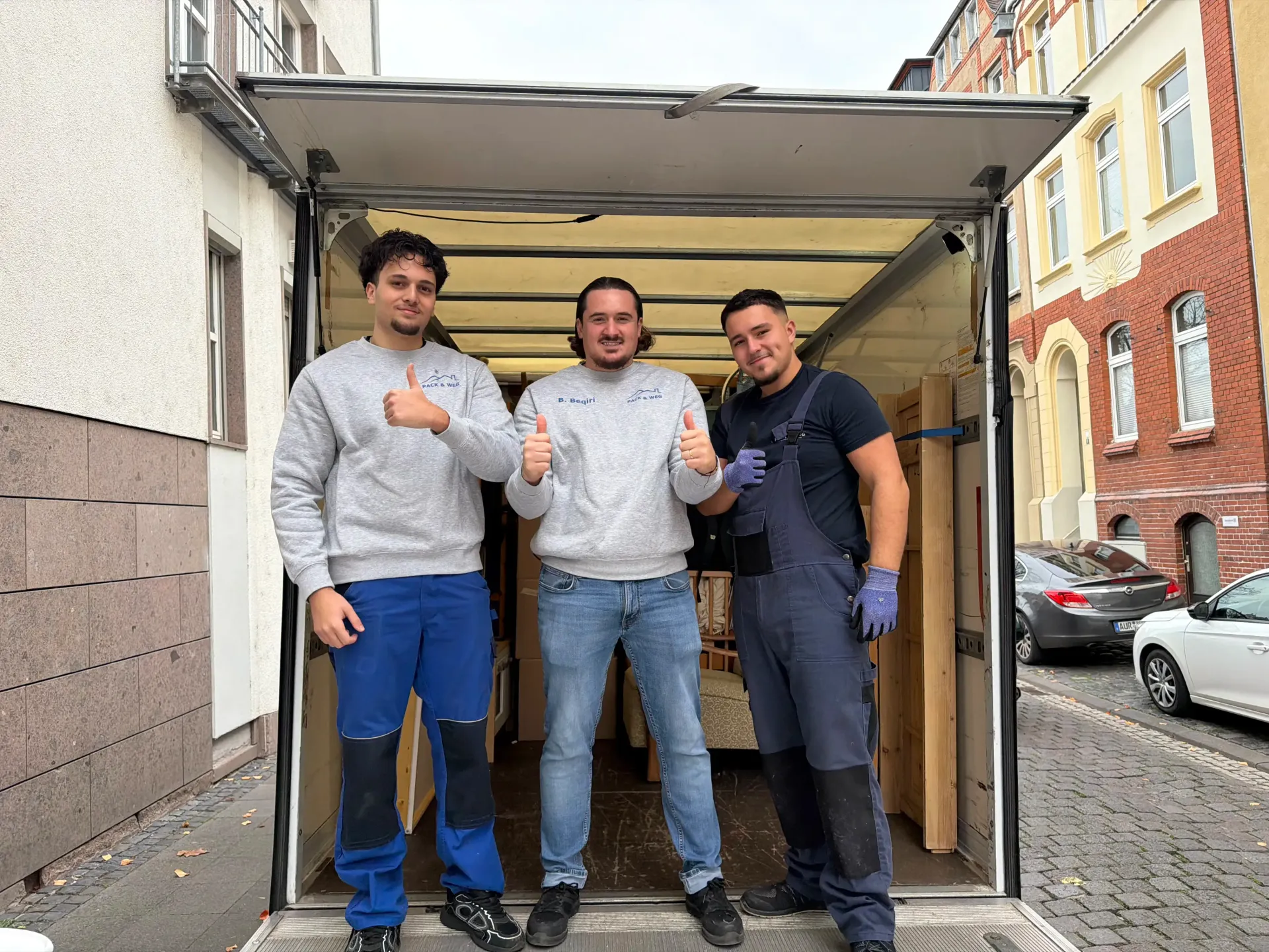 Drei Umzugshelfer stehen in einem offenen Umzugswagen und zeigen den Daumen nach oben. Im Hintergrund eine städtische Straße.