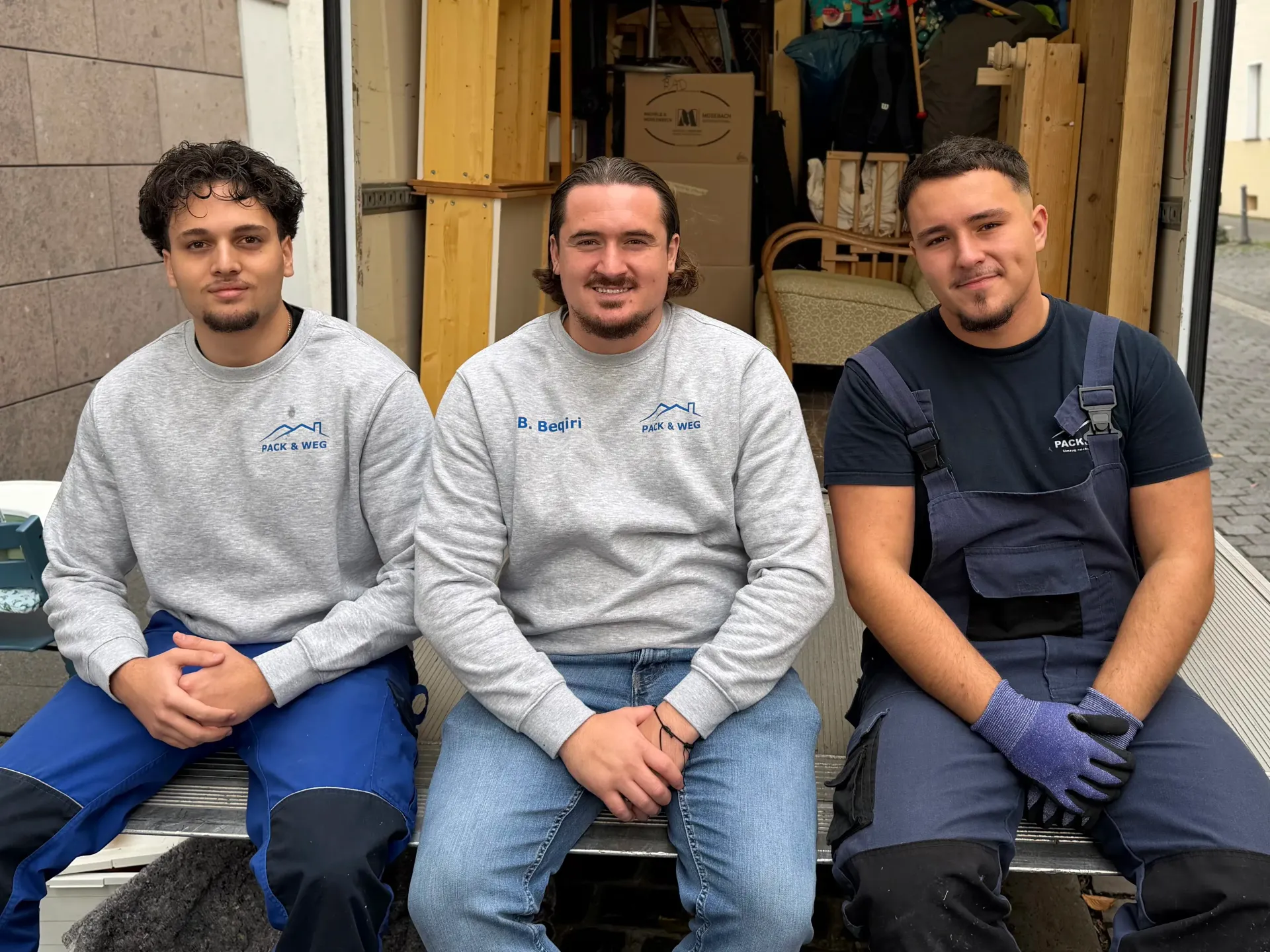 Drei Männer posieren neben einem Lieferwagen, zwei tragen graue Sweatshirts und blaue Hosen, einer trägt einen Overall und Handschuhe.
