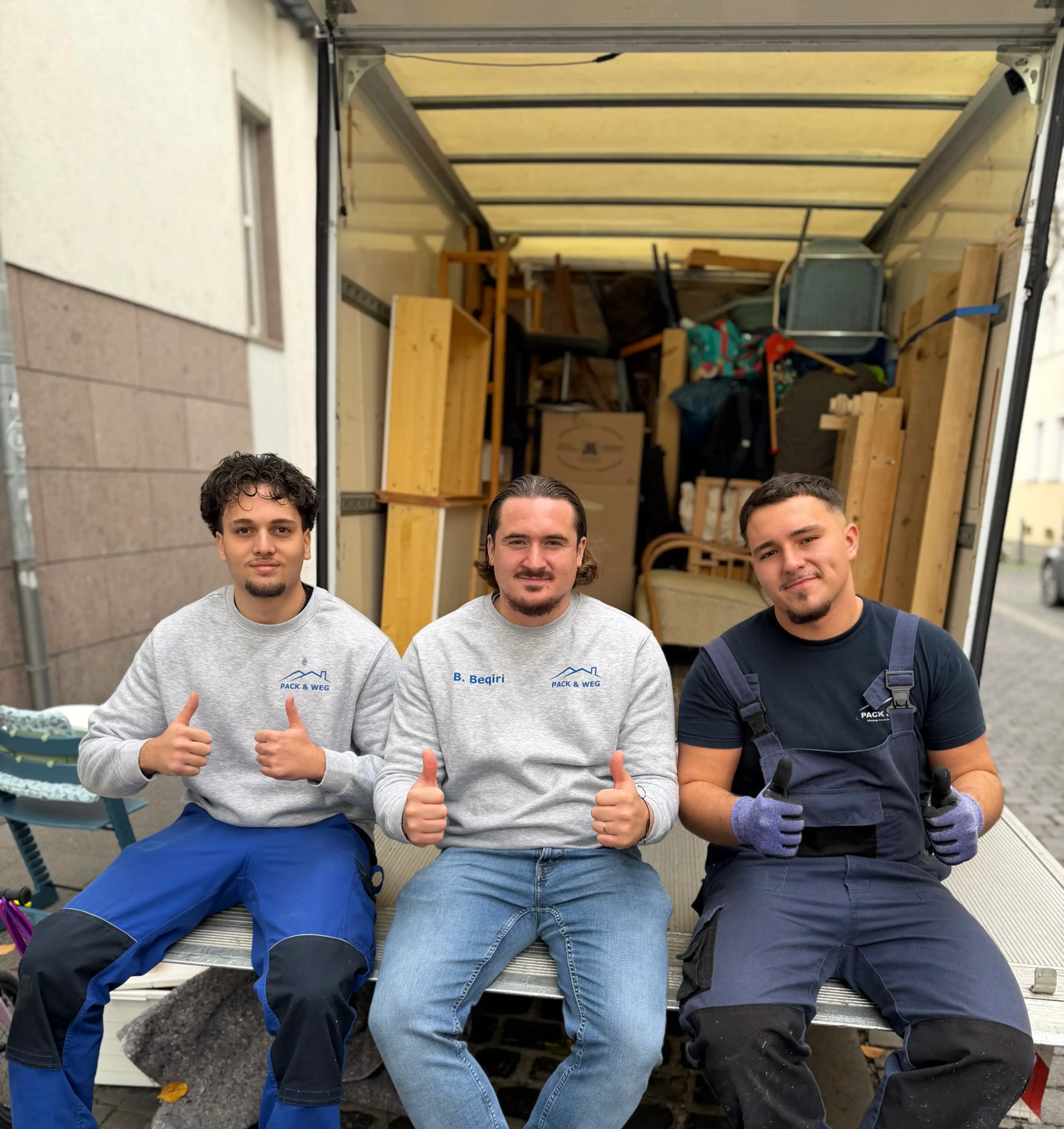 Drei Umzugshelfer mit erhobenen Daumen sitzen vor einem Umzugswagen, der mit Möbeln und Kisten beladen ist.