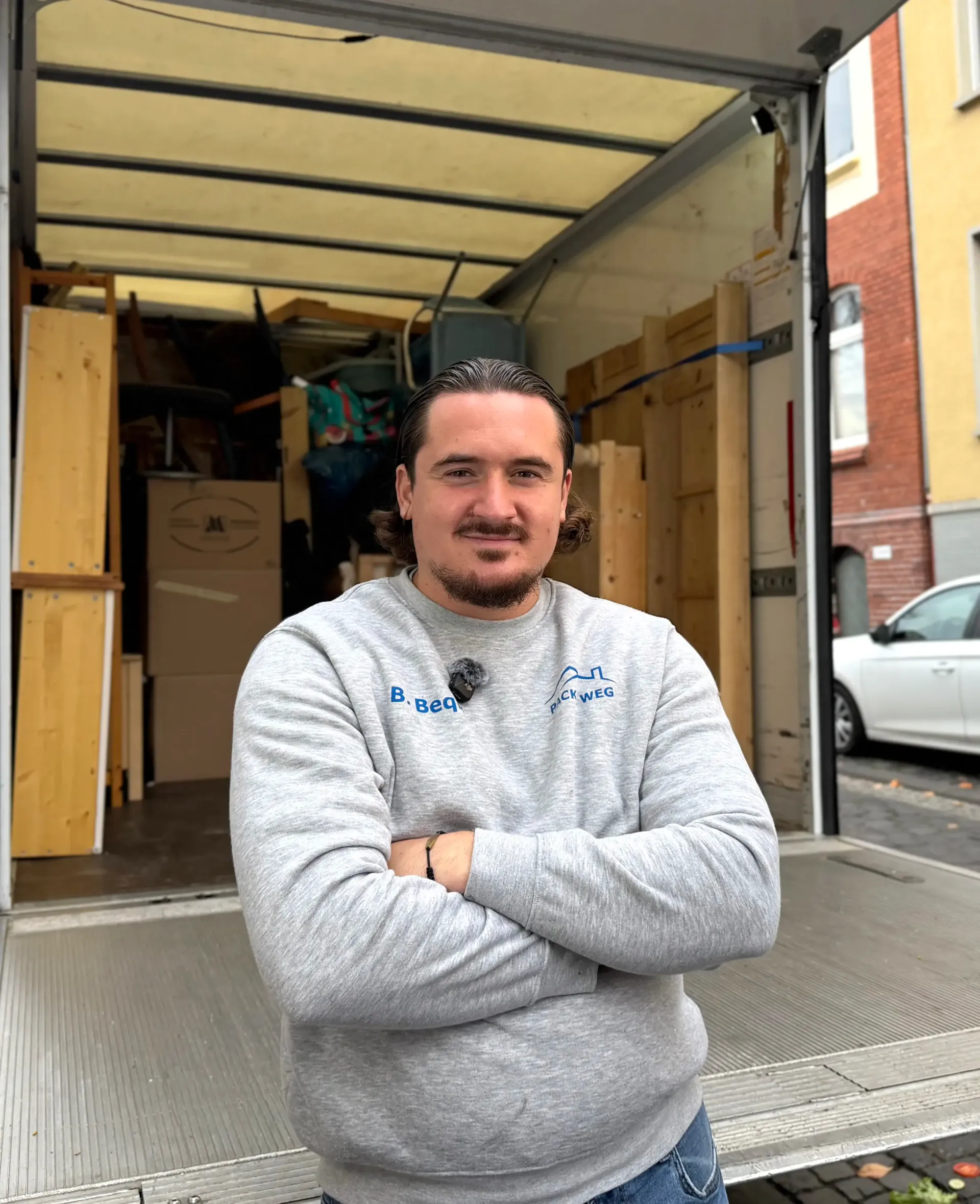 Ein Mann in grauem Sweatshirt, die Arme verschränkt, steht vor einem mit Möbeln beladenen Umzugswagen.